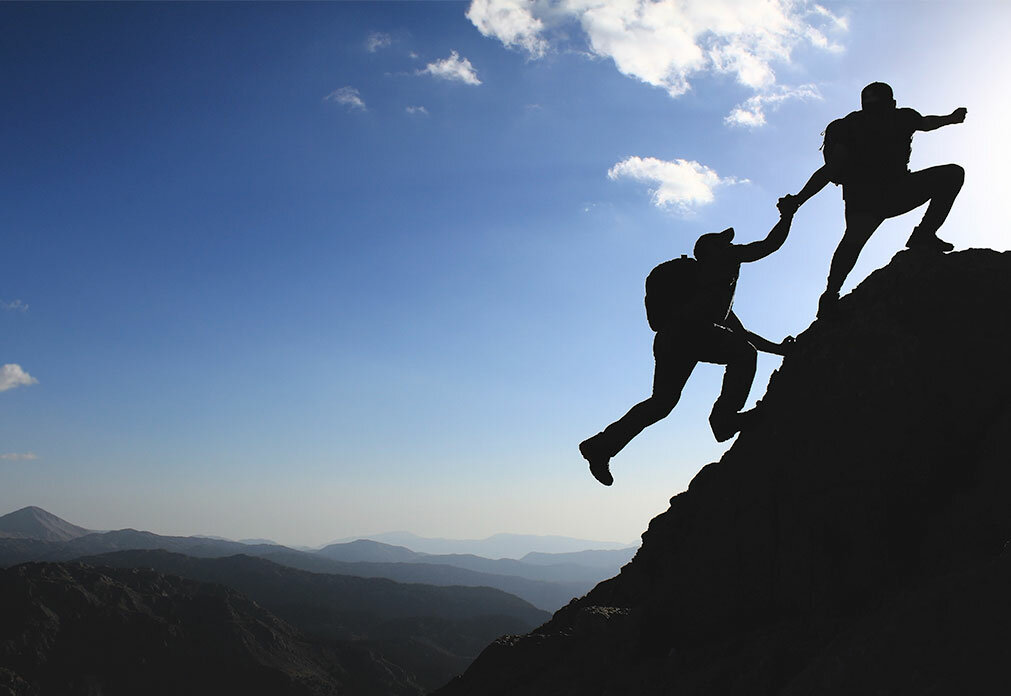 Two people climbing a mountain, one reaching out to help the other to the top