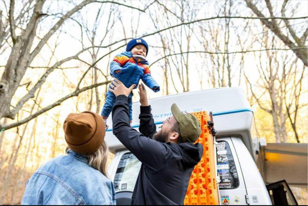 A young family throws their son up in the air as they all laugh outside of their Sprinter van in late fall in the woods of West Virginia.