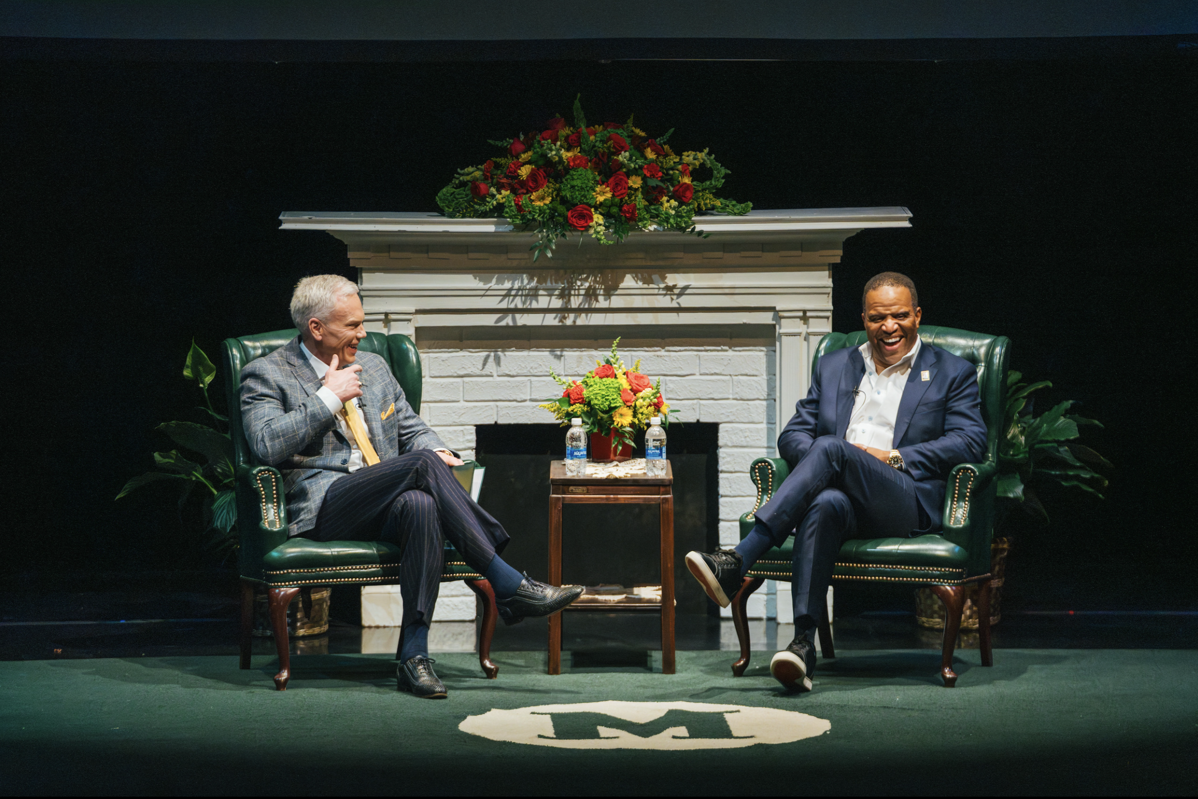 Brad D. Smith in a gray suit on stage with John Hope Bryant in a blue suit with a fireplace behind them
